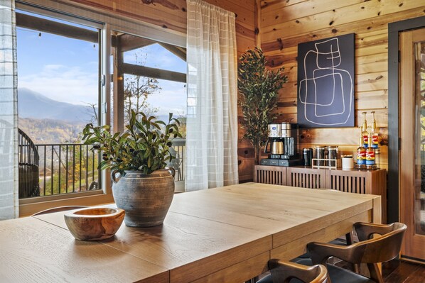 Ein Holzinterieur mit einem großen Tisch, einer Topfpflanze, einer Kaffeemaschine und Blick auf die Berge.
