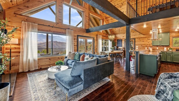 A spacious living room with a large sofa, a dining area, and a kitchen. There are large windows providing natural light and a view of the mountains. - Scenic Views•Hot Tub•Near Downtown (Gatlinburg)