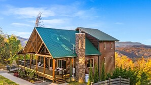 A log cabin with a green metal roof, a stone chimney, and a porch with outdoor seating. - Scenic Views•Hot Tub•Near Downtown (Gatlinburg)