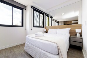 Une chambre d’hôtel moderne, dotée d’un grand lit, d’une table de chevet avec une lampe et offrant une vue sur la ville par les fenêtres.