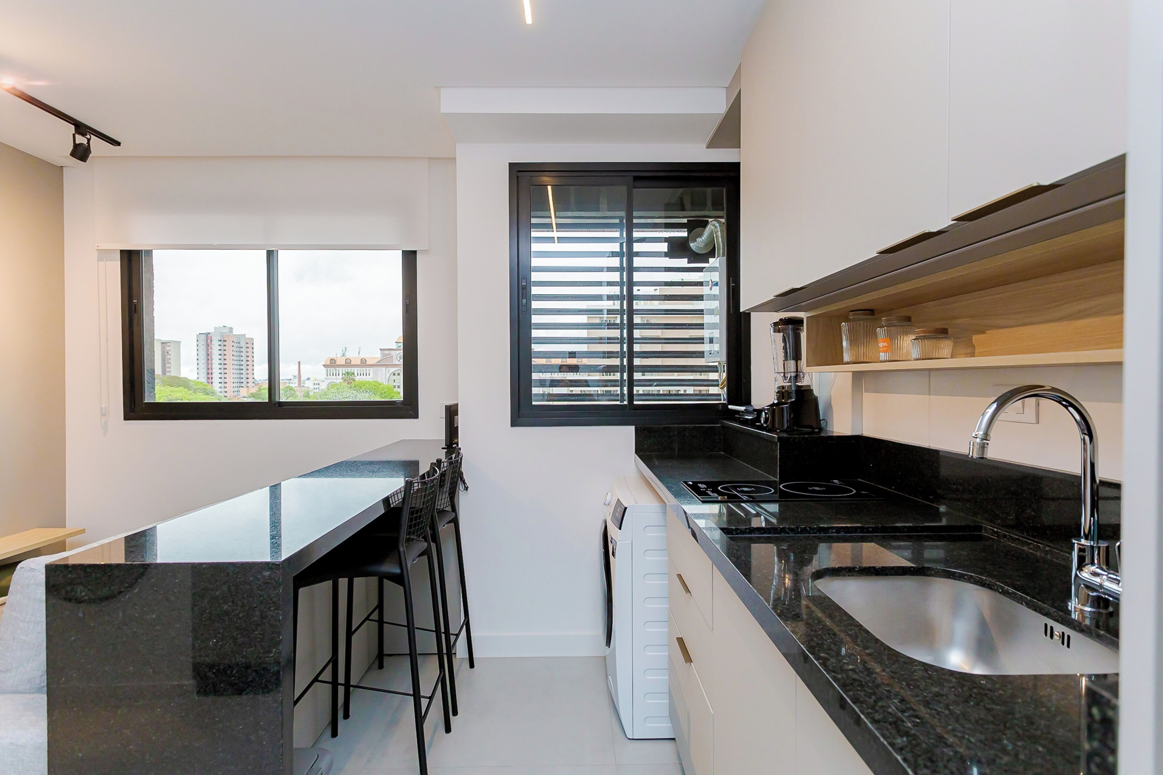 Een moderne keuken met een zwart aanrecht, een roestvrijstalen spoelbak en een inbouwoven. Er is een raam met zonwering en een uitzicht op gebouwen.