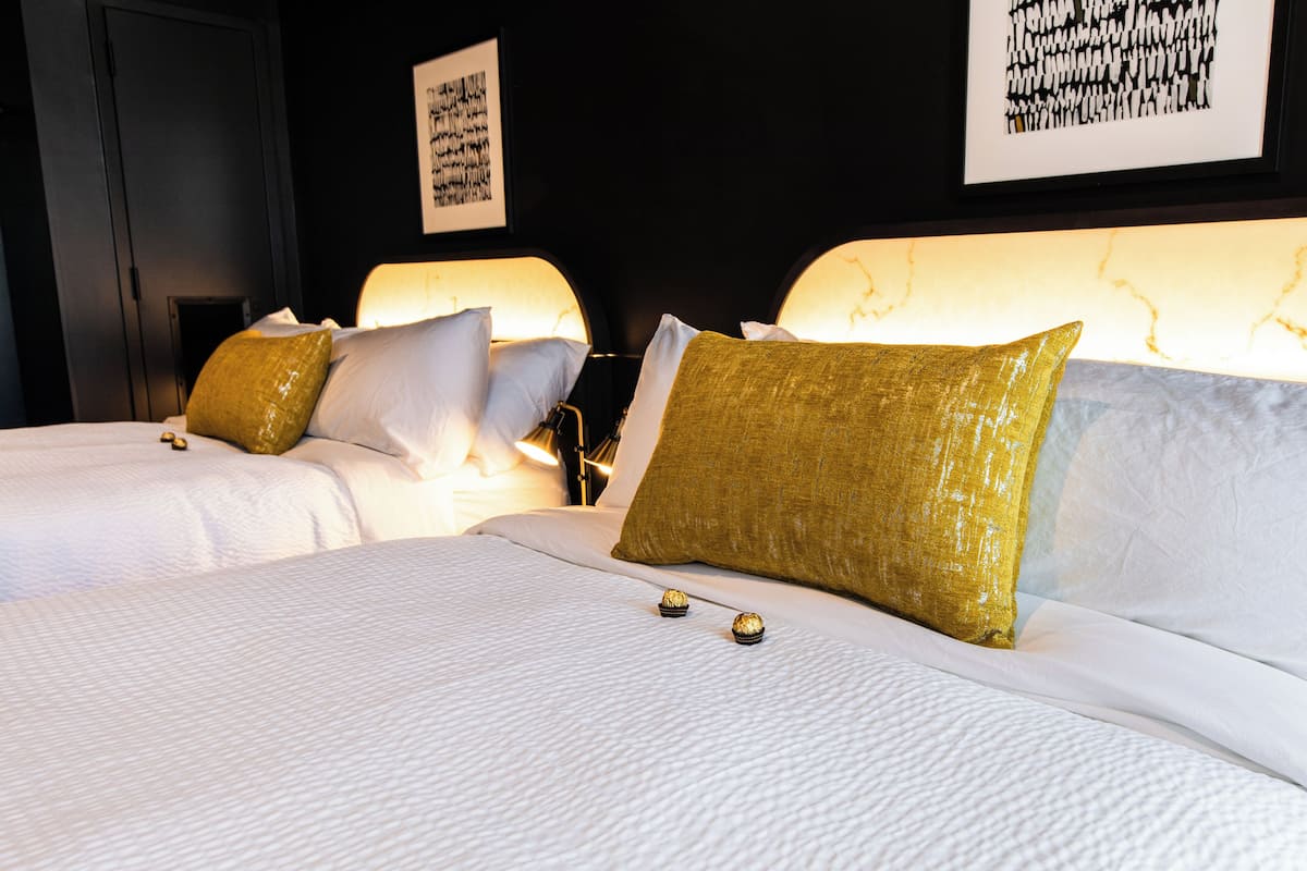Habitación de hotel con dos camas, cada una con ropa blanca y almohadas doradas.