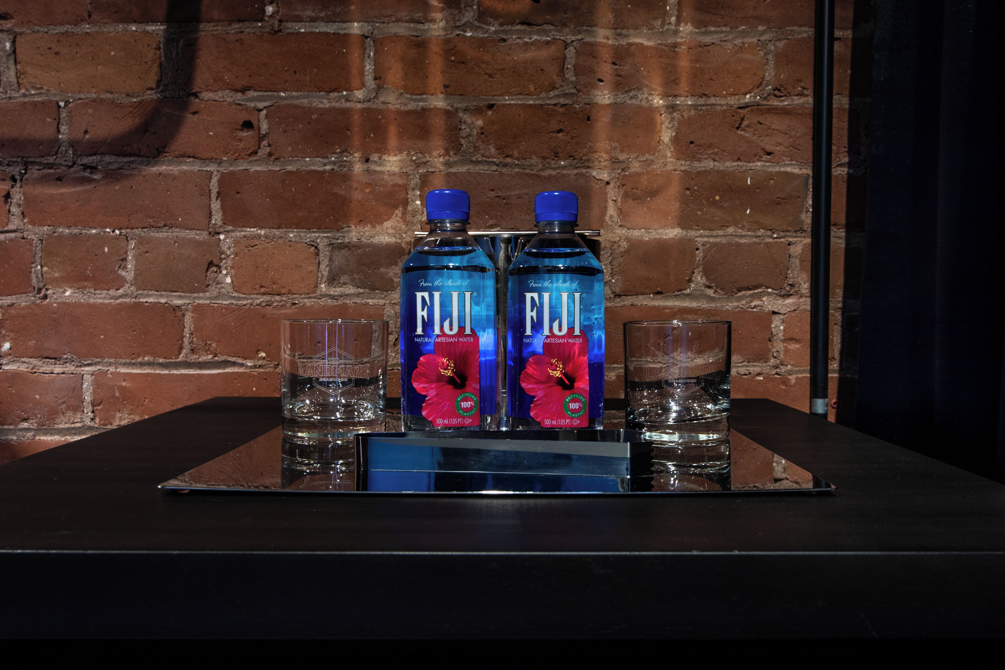 Deux bouteilles d’eau Fiji et deux verres sur un plateau, le tout posé contre un mur de briques.