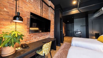 A modern hotel room with a brick wall, a flat-screen TV, a desk with a chair, a bed, and a potted plant.