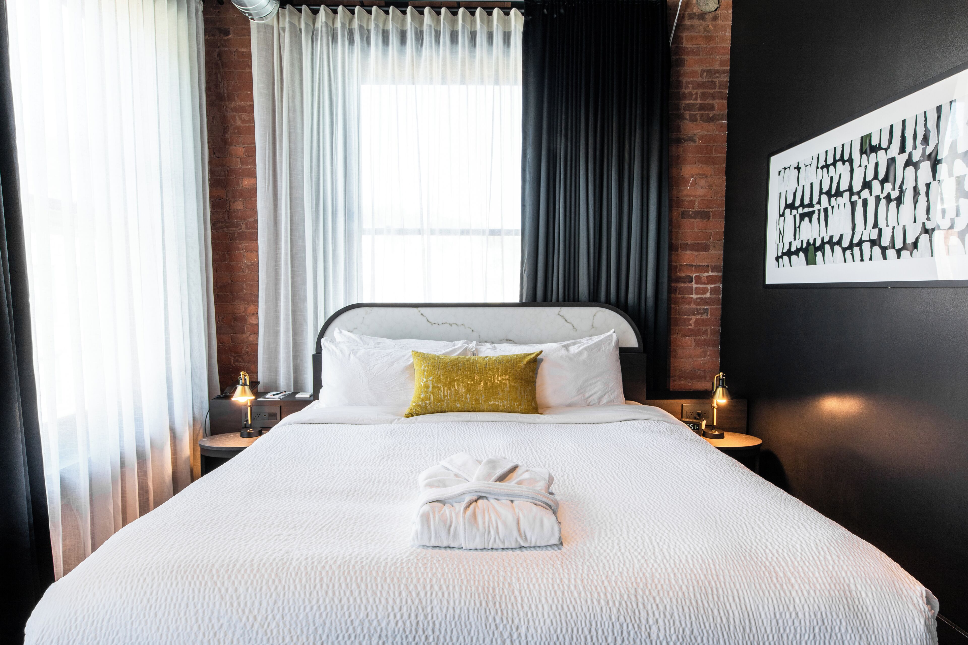 Een hotelkamer met een groot bed, een witte deken, een goudkleurig sierkussen, een netjes opgevouwen handdoek en een zwart-wit abstract schilderij aan de muur.