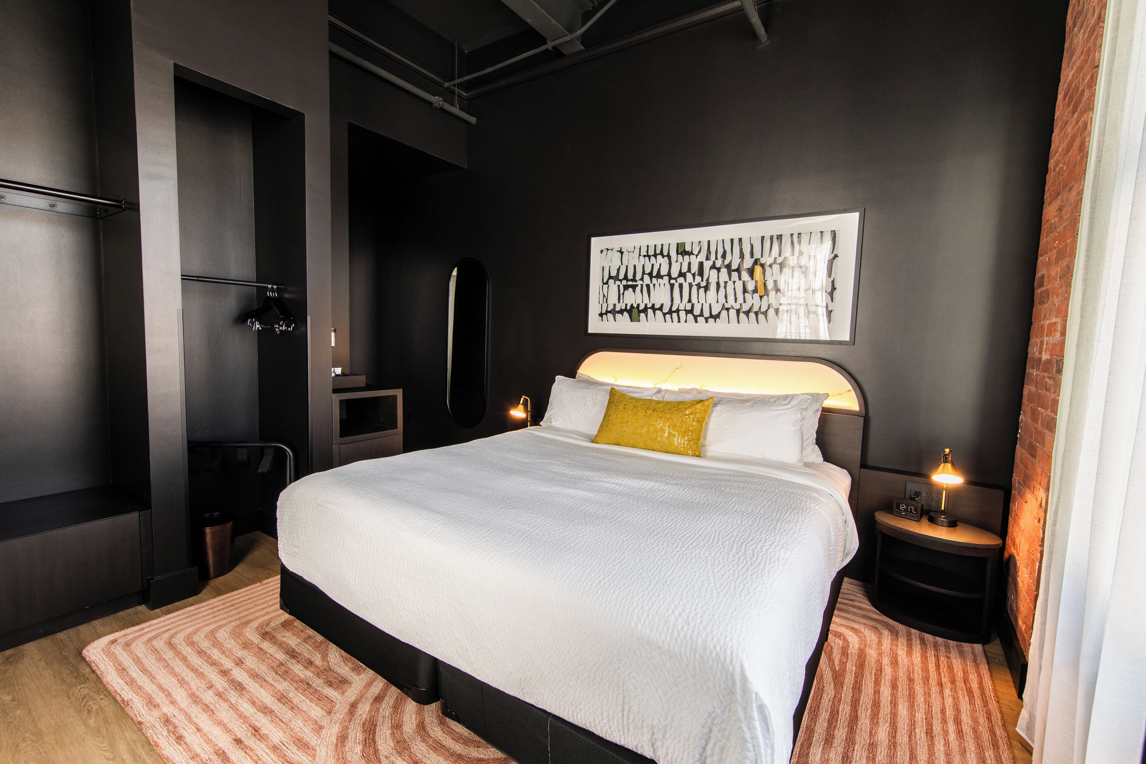 Une chambre moderne avec un grand lit, une table de chevet avec une lampe, une œuvre d’art accrochée au mur et une armoire de couleur sombre.