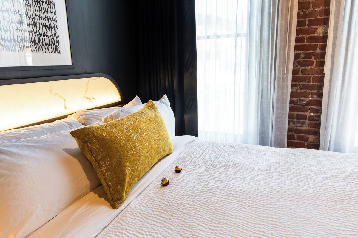 Una cama bien hecha con sábanas blancas y una única almohada color mostaza.