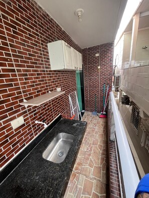 A narrow kitchen with a black granite countertop, a stainless steel sink, and a brick backsplash. There are white cabinets above the countertop and a green door in the background. - Flat 201 proximo centro Porto de Galinha (Ipojuca)