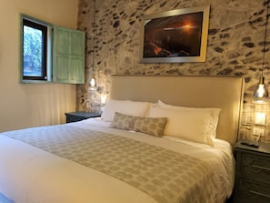 A bedroom with a stone wall, a bed with patterned bedding, a nightstand with a lamp, and a window with green shutters.