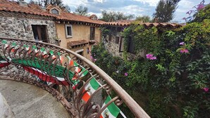 Un bâtiment en pierre avec un toit en tuiles, une balustrade en fer forgé ornée de drapeaux et un jardin planté de fleurs.