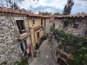 A narrow cobblestone street between stone buildings with terracotta roofs, a flag, and flowering vines.