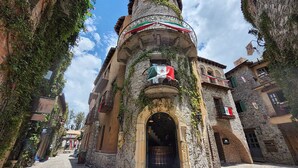 A narrow cobblestone street flanked by historic stone buildings with balconies, some adorned with flags.