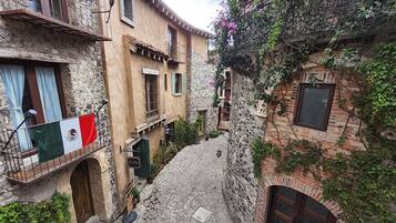 Une ruelle étroite pavée, bordée de bâtiments en pierre dotés de balcons, d’un drapeau et de plantes en pot.
