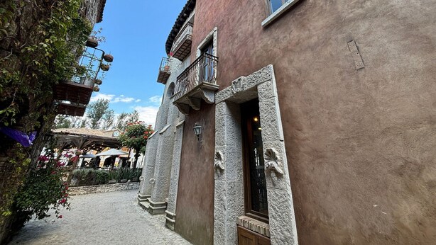 Une ruelle étroite bordée d’un immeuble caractérisé par des colonnes en pierre, une porte en bois et un balcon doté de rampes en métal.