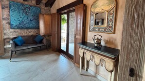 Une pièce avec un mur en pierre, un plafond en bois, un canapé avec des coussins bleus, une armoire avec un miroir et une porte donnant sur l’extérieur.