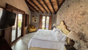 Une chambre avec un mur en pierre, un plafond en bois, un lit avec du linge de lit blanc, une télévision et un fauteuil.