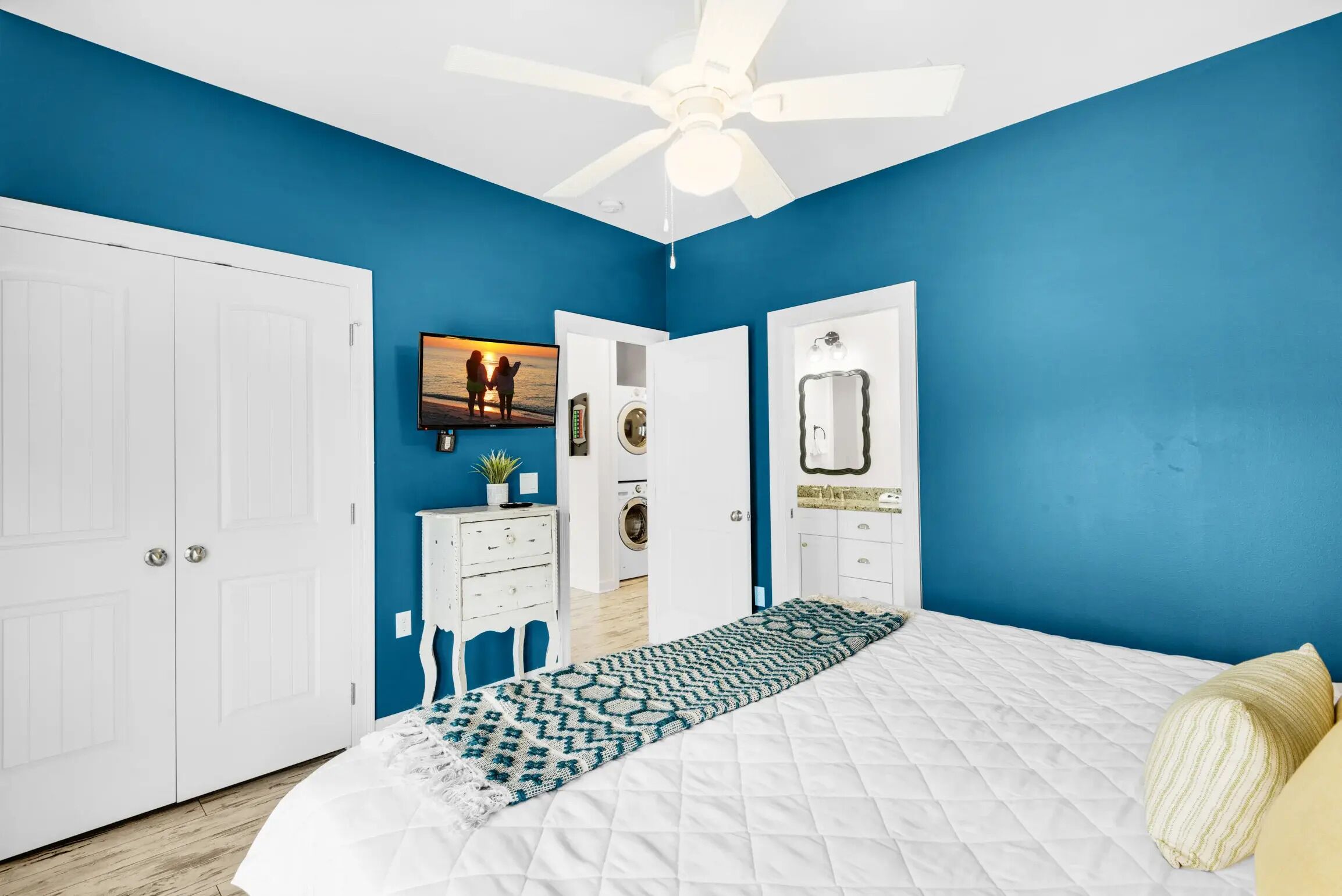 Un dormitorio con una cama blanca, ventilador de techo, pared azul y armario blanco.