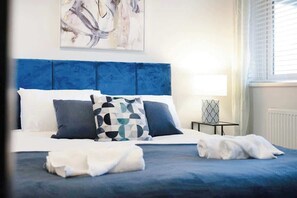 A bed with a blue headboard, white and patterned pillows, and a white blanket. - Modern 2 Bedroom Home With TV, WiFi, Parking (Hetton-le-Hole)