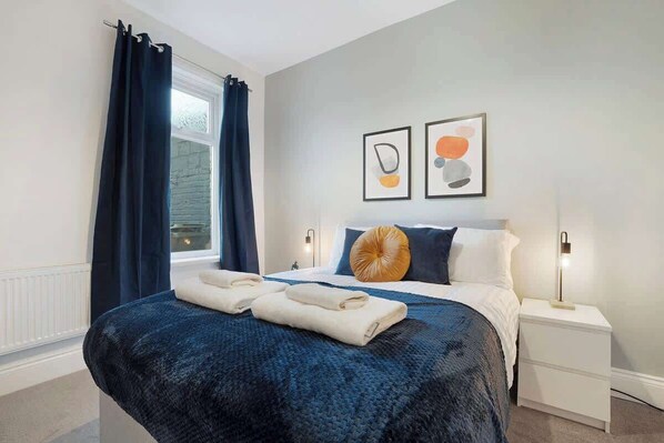 Une chambre à coucher avec un lit, deux tables de chevet, une fenêtre avec des rideaux et deux tableaux encadrés accrochés au mur.