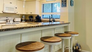 A modern kitchen with white cabinets, a granite countertop, and a bar area with stools. - 13Beach Stop Hilton Head (Hilton Head Island)