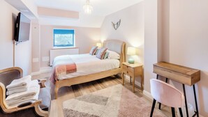 A bedroom with a bed, bedside table, lamp, chair, desk, TV, window, radiator, and a chandelier.