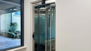 A modern elevator with glass doors, a visible emergency stop button, and a directional arrow sign.