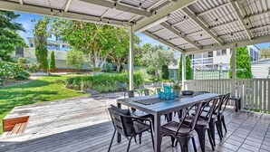 An outdoor patio with a table and chairs, surrounded by a wooden deck and a covered area with a metal roof. - Living The Dream Soldiers Point Home with BBQ (Soldiers Point)
