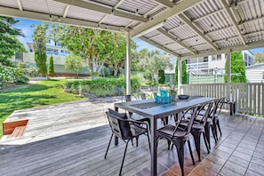 Een terras buiten, met een tafel en stoelen, omgeven door een houten terras en een overdekt gedeelte met een metalen dak.