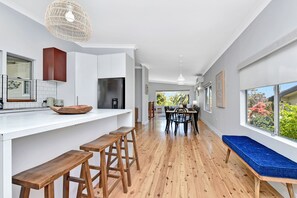 A modern kitchen with white cabinetry, a stainless steel refrigerator, wooden stools, and a dining area with a wooden table and chairs.
