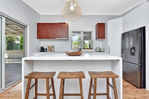 A modern kitchen with a central island, wooden stools, a stainless steel refrigerator, and a hanging light fixture. - Living The Dream Soldiers Point Home with BBQ (Soldiers Point)
