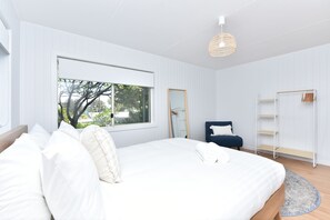 Ein Schlafzimmer mit großem Fenster, einem Bett mit weißen Leinentüchern, einem blauen Sessel, einem Kleiderschrank und einer Pendelleuchte.