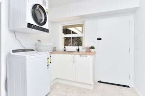 A laundry room with a front-loading washer and dryer, a window, and a countertop with a sink and storage containers. - Living The Dream Soldiers Point Home with BBQ (Soldiers Point)