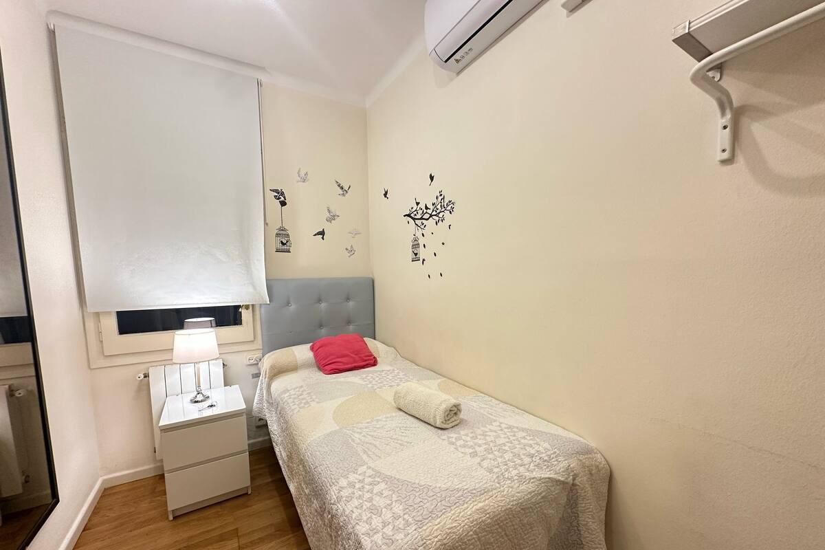Une petite chambre à coucher, bien rangée, avec un lit, une table de chevet, une lampe et un climatiseur fixé au mur.