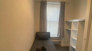 Een kleine, smalle kamer met een enkel bed, een raam met gordijnen en een inbouwkast met planken.