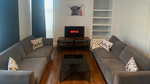 Een moderne woonkamer met twee grijze banken, een salontafel, een televisie waarop 'NETFLIX' staat en een kroonluchter.