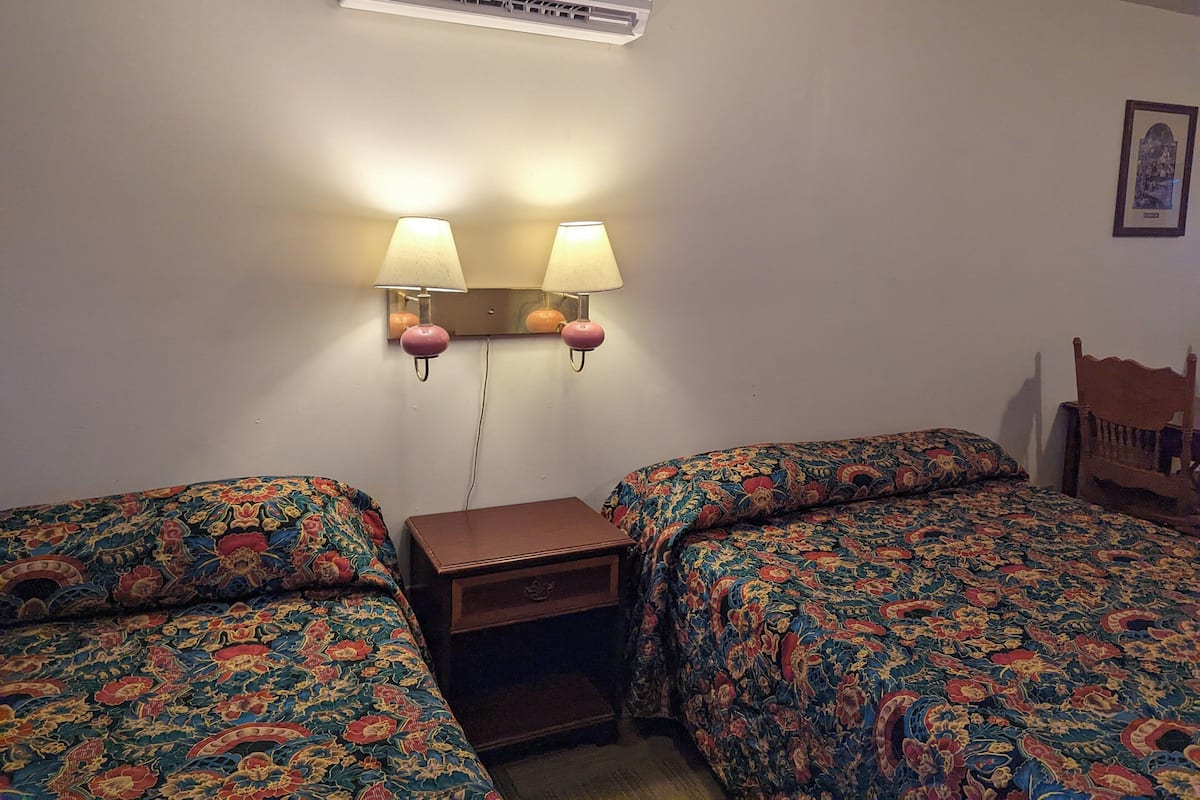 A hotel room with two beds, each with a floral-patterned bedspread, a nightstand with a lamp, and an air conditioning unit above the beds.