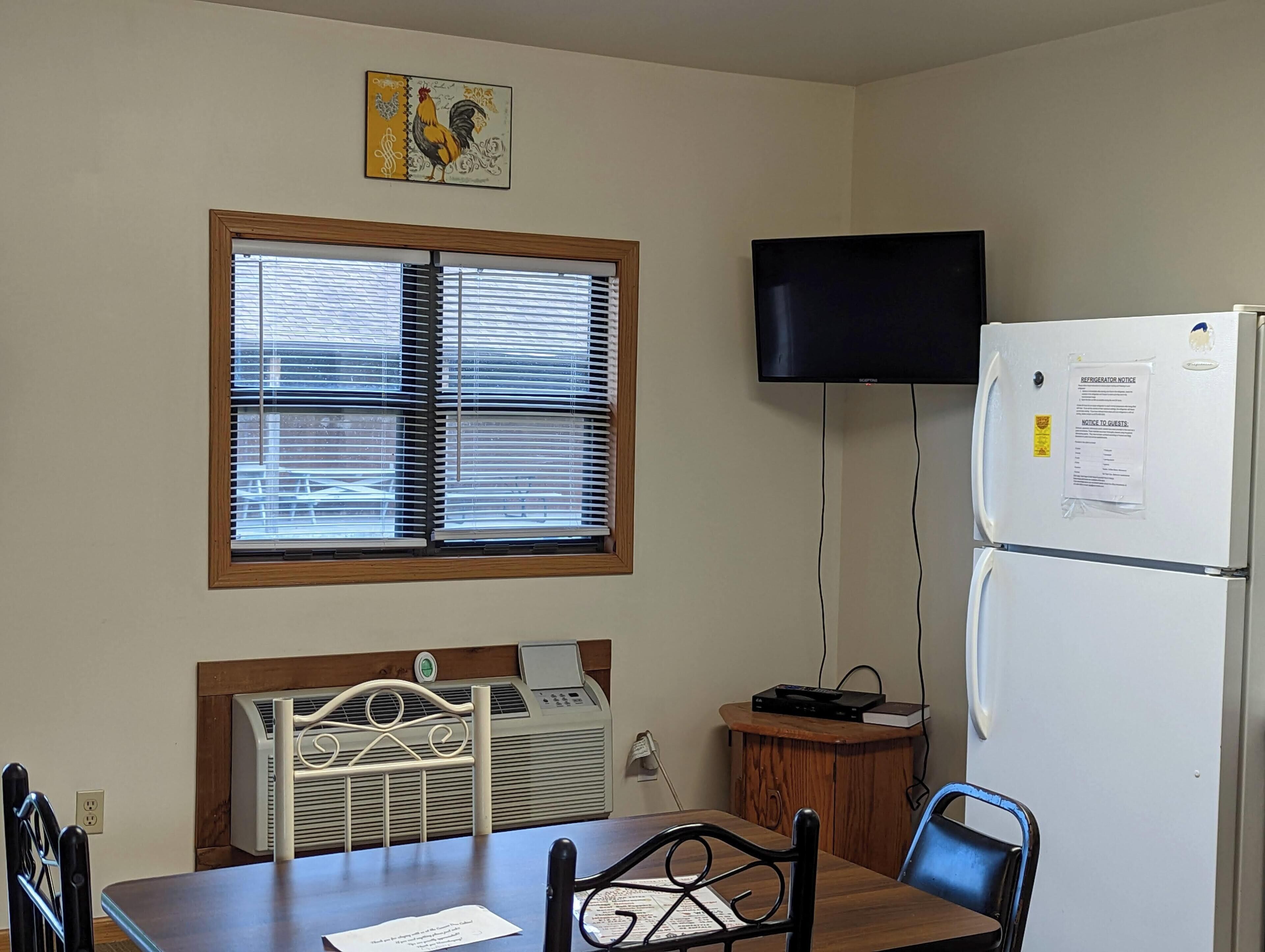 A small, well-lit room with a wooden table, chairs, a window with blinds, a wall-mounted TV, and a refrigerator with a notice.