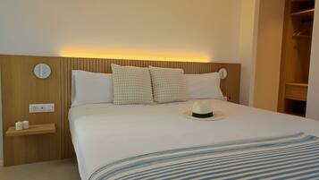 Una cama bien hecha con una colcha a rayas, dos almohadas y un sombrero sobre la cama. Se observa un cabecero de madera, una mesita de noche con dos velas y un enchufe de pared.