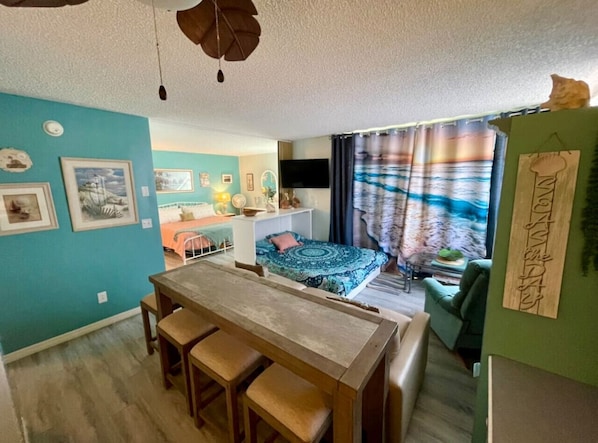 A compact living space with a dining area, a bed with a patterned bedspread, a green sofa, and a television. - Sunset & Ocean Front Views-FM-Beach(Pool is Open) (Fort Myers Beach)