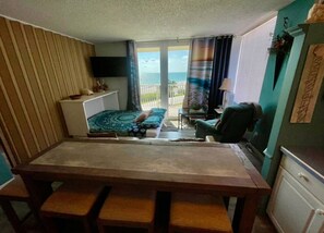 A dining area with a table and benches, a sofa, a TV, and a view of the ocean through a sliding door.
