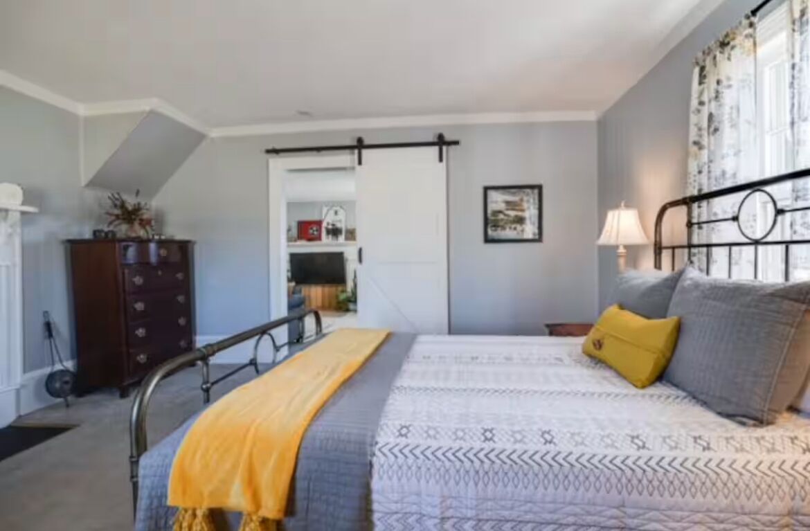 A bedroom with a bed, a nightstand, a lamp, a dresser, and a door leading to another room.