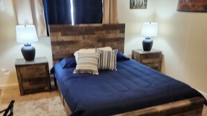 A bedroom with a wooden bed frame, two bedside tables with lamps, a blue bedspread, and a window with curtains. - Charming studio in enchanting Colorado Springs with WiFi, AC (Colorado Springs)