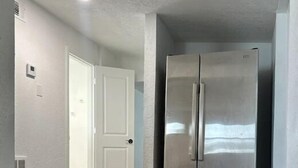 Ein Flur mit einem Edelstahlkühlschrank, einer weißen Tür und einem Lüftungsgitter an der Wand.