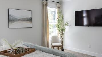 A bedroom with a bed, a chair, a potted plant, a flat-screen TV, and a window with curtains.