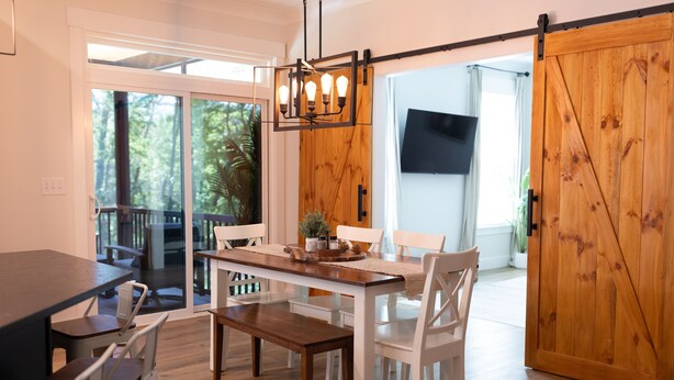 Un comedor con una mesa de madera, sillas blancas y un banco. Hay una puerta corrediza con acabado de madera y un televisor montado en la pared.