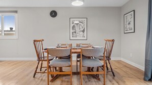 Un comedor con una mesa redonda, cuatro sillas de madera, un reloj y dos cuadros en la pared.
