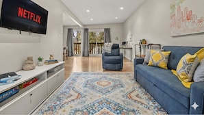 Una sala de estar moderna con un televisor de pantalla plana mostrando 'NETFLIX', un sofá azul con cojines amarillos y grises, una alfombra estampada y un cuadro con una vista urbana en la pared.