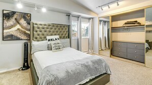 Een slaapkamer met een gestoffeerd hoofdeinde, een bed met grijze beddengoed, een nachtkastje, een elektrische kachel, een groot gekaderd kunstwerk, een kast met spiegel en een raam met gordijnen.