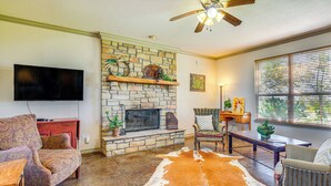 A living room with a stone fireplace, a ceiling fan, a television mounted on the wall, and a large cowhide rug. - 30 Mi to Fort Worth: Rural Retreat w/ Patio! (Godley)