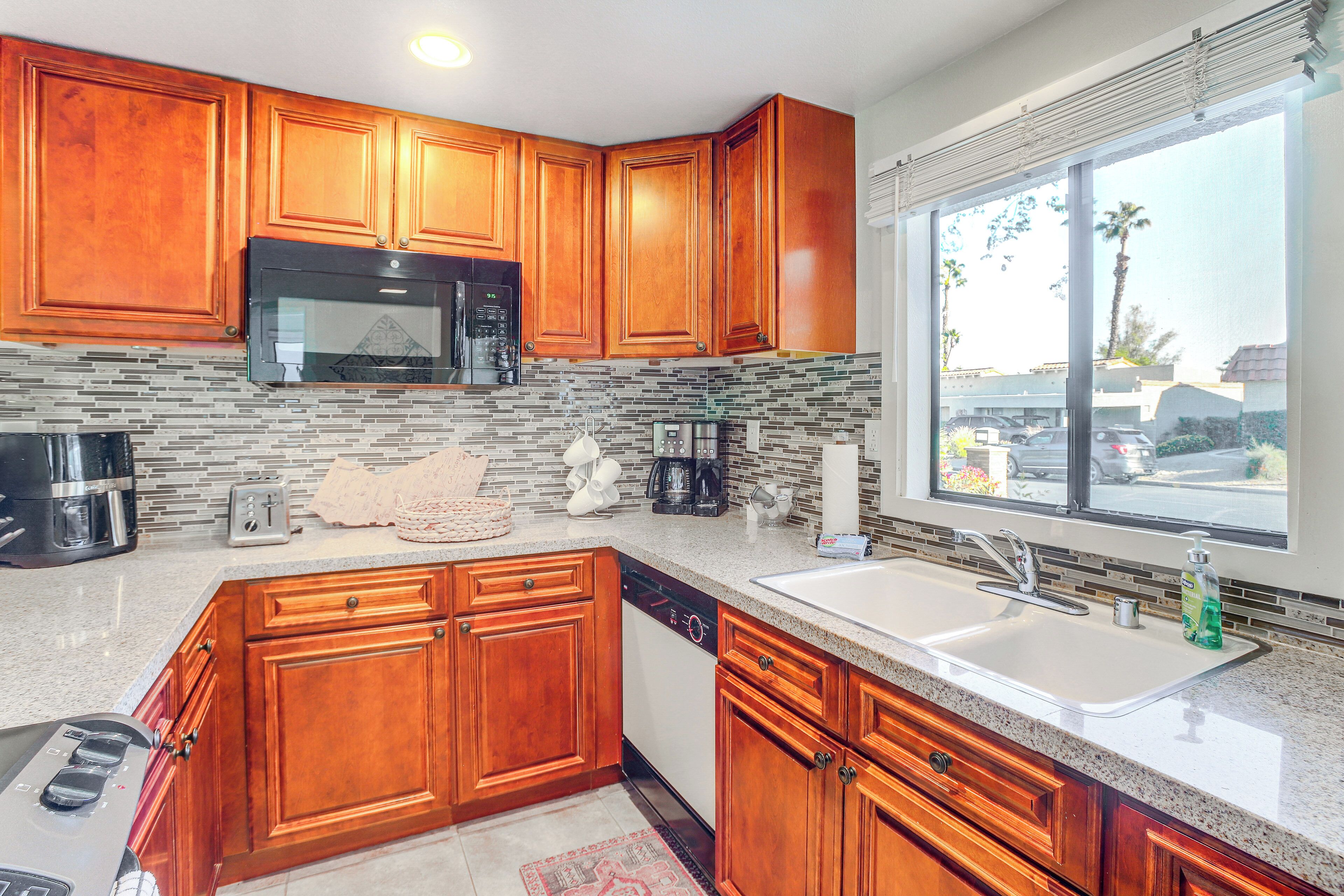 A kitchen with wooden cabinets, a stainless steel microwave, a dishwasher, and a window with a view of palm trees and parked cars.